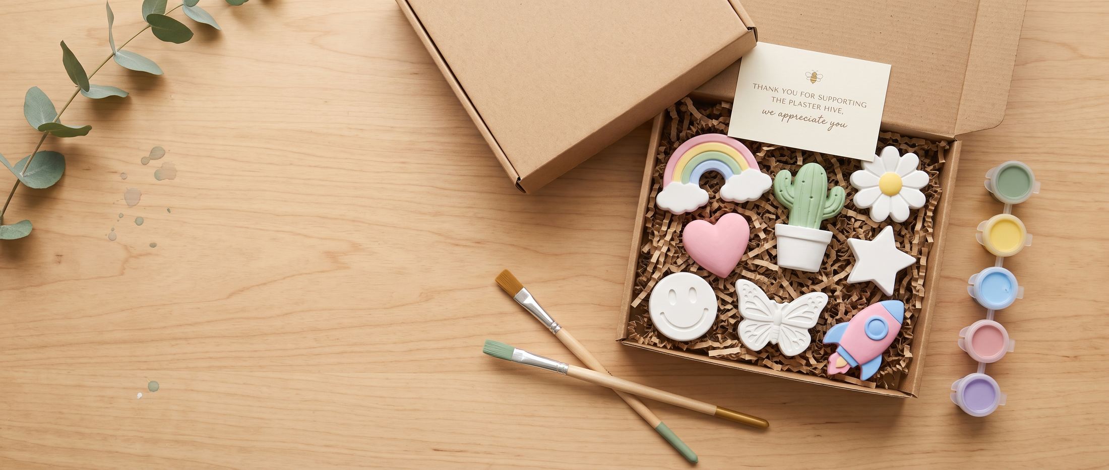 Wide flat-lay of a wood table with an open Plaster Hive kit box of painted plaster shapes, paintbrushes, pastel paint jars, and eucalyptus greenery.